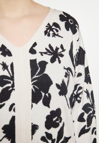 Sweter dzianinowy w kolorze kremowym z dekoltem w serek, zdobiony czarnym wzorem kwiatowym. Materiał wydaje się miękki i ma fakturowane wykończenie.