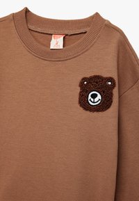 Bruine sweatshirt met een ronde halslijn met een geborduurde berengezicht in donkerbruin en wit, gemaakt van zacht materiaal met een gladde textuur.