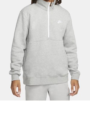 Osoba w jasnoszarej półrozpinanej bluzie Nike i dopasowanych jasnoszarych spodniach z logo Nike na lewej piersi i lewym udzie.
