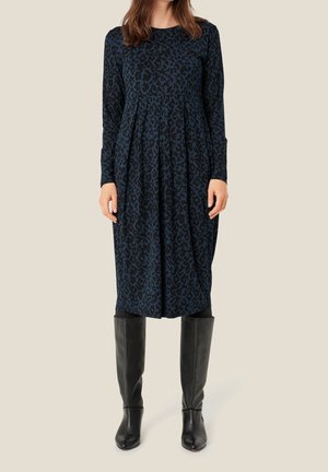 Femme portant une robe bleu foncé à manches longues, longueur genou, avec un imprimé léopard noir et des bottes en cuir noires montant jusqu'aux genoux.
