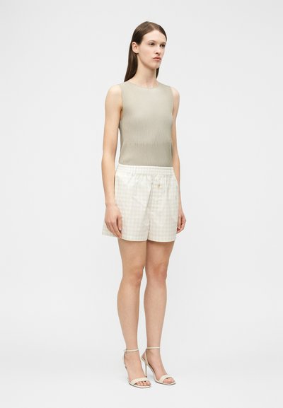 Νεαρή γυναίκα όρθια, φορούσε αμάνικο ριμπ μπεζ τοπ, φαρδιά σορτς με ψηλή μέση και ανοιχτόχρωμο καρό μοτίβο, και μπεζ πέδιλα με ψηλό τακούνι.