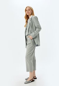 Traje entallado de color gris claro con blazer estructurado y pantalones cortos. Tejido texturizado, camisa blanca debajo y zapatos de tacón tipo slingback negros.