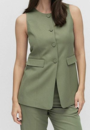 Kvinde iført ærmeløs olivengrøn knappet vest med klaplommer og matchende bukser, stående mod en ensfarvet lys baggrund.