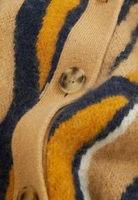 Ullkofta med djärva ränder i gult, marinblått och krämvit. Texturerat tyg med lätt känsla och en tydlig, rund knappstängning.
