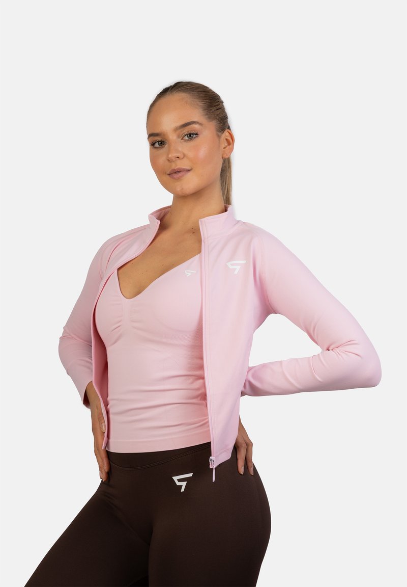Lys pink atletisk zip-up jakke med høj krave, tætsiddende design og hvidt logo. Bæres over en matchende pink top og brune leggings.