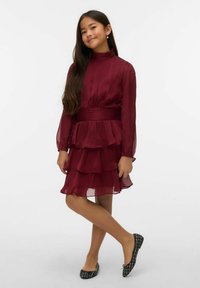 Vestito bordeaux a balze con maniche lunghe di tessuto trasparente, collo alto e cintura testurizzata. Abbinato a ballerine nere con motivo a fantasie.