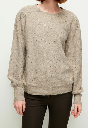 Femme portant un pull en maille beige avec de subtils éclats métalliques et des manches longues bouffantes, associé à un pantalon marron foncé.