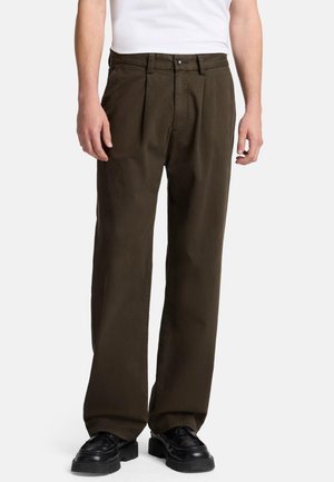 Dunkelolivgrüne Hose aus leichtem Stoff, mit Falten, einem Bund mit Gürtelschlaufen und einem Knopfverschluss, kombiniert mit schwarzen Schuhen.