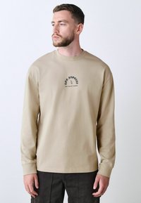 Beige langærmet t-shirt i bomuld med rund halsudskæring og et lille trykt design på brystet med tekst og grafiske elementer.