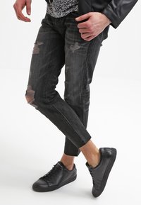 Jeans de mezclilla negra con detalles rasgados, diseño slim fit y una textura desgastada, combinados con zapatillas negras de corte bajo.