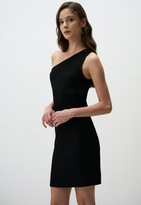 Abito nero a costine con design a spalla unica, silhouette aderente, texture liscia e lunghezza sopra il ginocchio. Nessun motivo visibile o accessori.