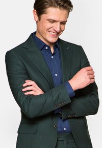 Ciemnozielona, dopasowana marynarka o teksturowanym wykończeniu, noszona na niebieskiej koszuli z guzikami. Wyraziste guziki na mankietach i kołnierzu.