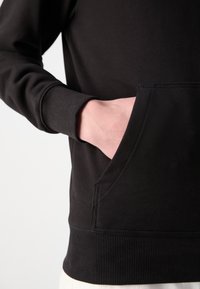 Hanorac negru cu tiv și mansete riglate, având o buzunar frontal și o textură netedă, arătând o parte a unei mâini care se îndreaptă spre buzunar.