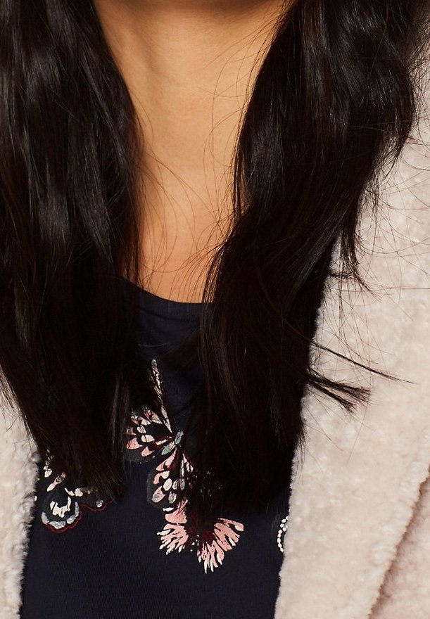 Cheveux longs noirs sur une chemise bleu marine avec un motif de papillon rose et blanc, et une veste polaire crème portée sur les épaules.