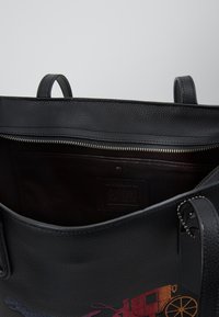 Sort struktureret lædertaske med lynlåslukning øverst, der har en dyb indvendig lomme og et præget mærkat på siden.