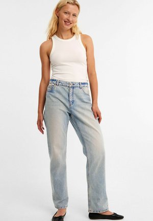 Jean en denim délavé avec une coupe ample, présentant une taille haute, cinq poches, et une couture bleue accentuée ; associé à un débardeur blanc à côtes.