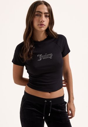 Jeune femme aux longs cheveux bruns portant un t-shirt court noir avec « Juicy » en strass et un pantalon noir à cordon, debout les mains derrière.