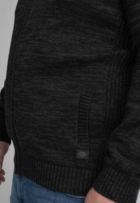 Suéter de punto gris oscuro con un patrón texturizado, puños y dobladillo acanalados, que presenta una pequeña etiqueta en el lado inferior izquierdo.