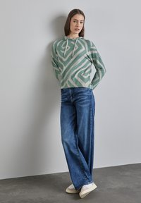 Sweat-shirt à capuche vert et beige avec un motif ondulé, associé à un jean en denim bleu ample et des baskets de couleur claire.