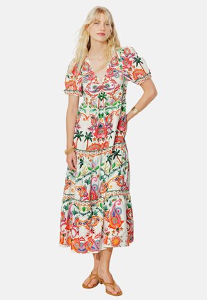 Femme portant une robe midi colorée à motifs floraux et paisley, manches bouffantes et sandales beige, debout avec une main sur la hanche.