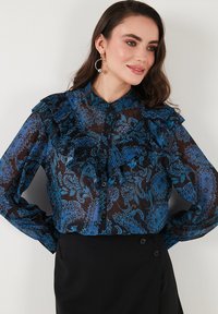 Blue and black sheer blouse with a paisley pattern, features ruffles along the shoulders and a button-up front, paired with a black skirt.
