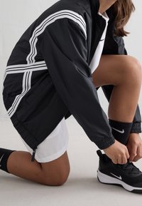 Person kneeling ties black Nike sneakers, wearing black jacket with white stripes, white shorts, and black Nike socks on a white floor.