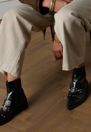 Stivaletti neri in pelle con borchie dorate e dettagli di frange, caratterizzati da una punta a punta. Indossati con pantaloni beige chiaro a gamba larga.