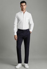 Chemise blanche à manches longues avec une poche, associée à un pantalon bleu marine et des baskets blanches, se tenant devant un fond gris neutre.