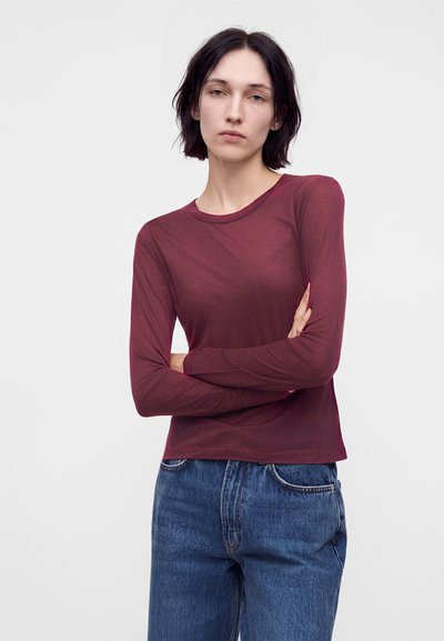 Jeune femme aux cheveux courts et foncés, portant une chemise bordeaux à manches longues et un jean bleu, debout, les bras croisés, devant un fond uni.