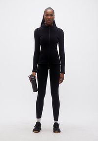 Γυναίκα με μαύρη αθλητική ενδυμασία που κρατά ένα μαύρο μπουκάλι νερού, στέκεται μπροστά σε λευκό απλό φόντο.