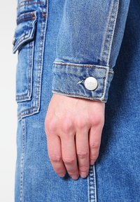 Sininen farkkutakin hiha, jossa on hopeinen nappidetail. Kankaassa on teksturoitu kudonta ja näkyvät ompelulinjat, jotka korostavat muotoilua.