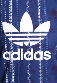 Blå mönstrad tyg har vertikala vita randdetaljer, med en framträdande vit Adidas-logotyp i mitten.