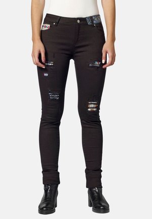 Úzké černé džíny s rozervanými nášivkami a barevnými látkovými detaily, nošené s černými kotníkovými botami na podpatku a bílým zašitým topem.