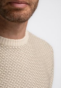 Beige knitted sweater with textured pattern and ribbed crew neck. Close-up view focuses on fabric details and neckline.