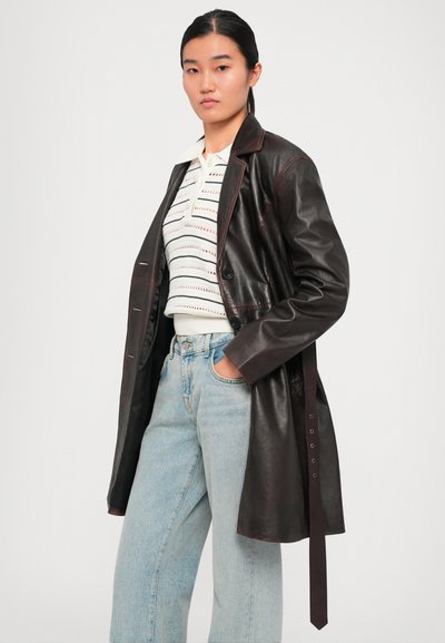 Giovane donna che indossa un cappotto di pelle marrone scuro, un top lavorato a maglia bianco e jeans azzurri a vita alta, in piedi con una mano in tasca.