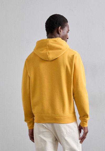 Felpa gialla con cappuccio, dal tessuto morbido, con polsini a costine e vestibilità rilassata, indossata sopra pantaloni chiari. Vista posteriore.