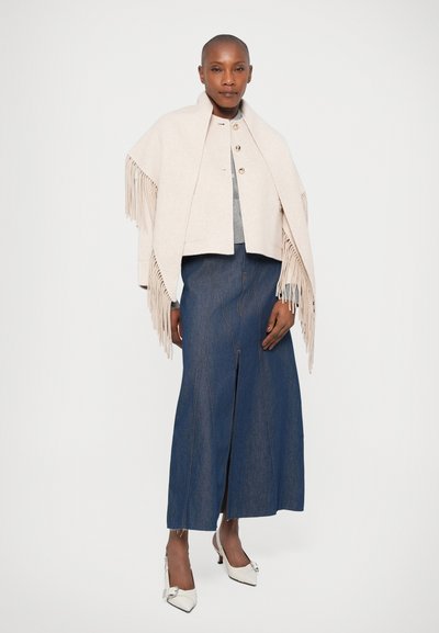 Beige kort poncho med fransdetaljer, bärs över en grå topp och en mörkblå denim-maxikjol med en frontslits och sömmar. Vita klackskor.