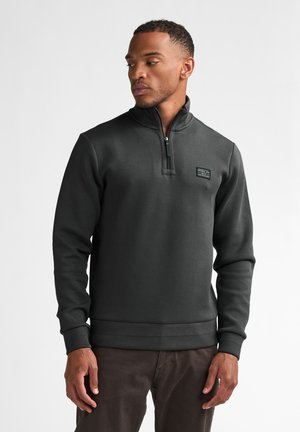 Man wearing a dark gray long-sleeve quarter-zip sweatshirt with a small logo patch on the left chest, standing against a plain white background.