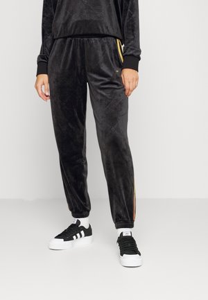 Person wearing black velour jogger pants with yellow side stripes and black-and-white Adidas sneakers, standing against a plain background.