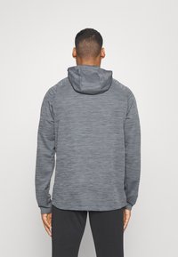 Uomo che indossa una giacca con cappuccio grigia e testurizzata e pantaloni neri, in piedi rivolto verso uno sfondo chiaro uniforme con le braccia rilassate lungo i fianchi.