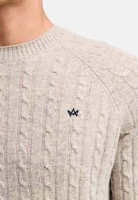 Beżowy sweter w splot kablowy z prążkowanym okrągłym dekoltem i małym czarnym emblematem na piersi. Tekstura wydaje się miękka i ciepła.