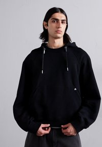 Črna kapuca z vrvicami, ki ima kenguru žep in rebraste manšete. Material se zdi mehak in teksturiran, s sproščenim krojem.