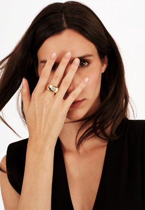 Goldring mit einem auffälligen klaren Stein, glatte Oberfläche, ovale Form, auf dem Finger eines Models, teilweise von ihrer Hand verdeckt.