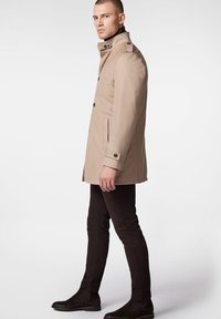 Beżowy trencz z wysokim kołnierzem, dwiema przednimi kieszeniami i guzikami. Model w czarnych spodniach i botkach na kostkę, stojący z boku.