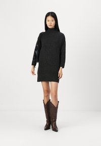 Robe noire en tricot avec un col haut et des manches longues, associée à des bottes de cowboy marron ornées d'un motif. Modèle tenant un sac à main noir.