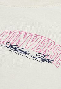 Sweatshirt beige avec un texte rose et bleu marine en relief indiquant "CONVERSE Athletic Dept." avec un design classique courbé. Tissu texturé.
