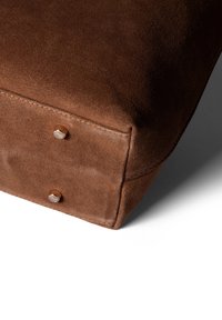 Coussin en daim brun avec une texture lisse et des bords arrondis, présentant deux accents métalliques sur le coin pour un détail supplémentaire.