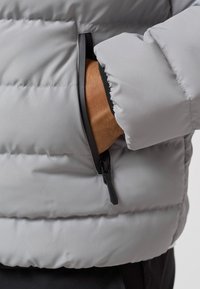 Giacca a vento grigia con imbottitura, dotata di tasca con zip e tessuto testurizzato, con cuciture visibili e un leggero finish lucido.
