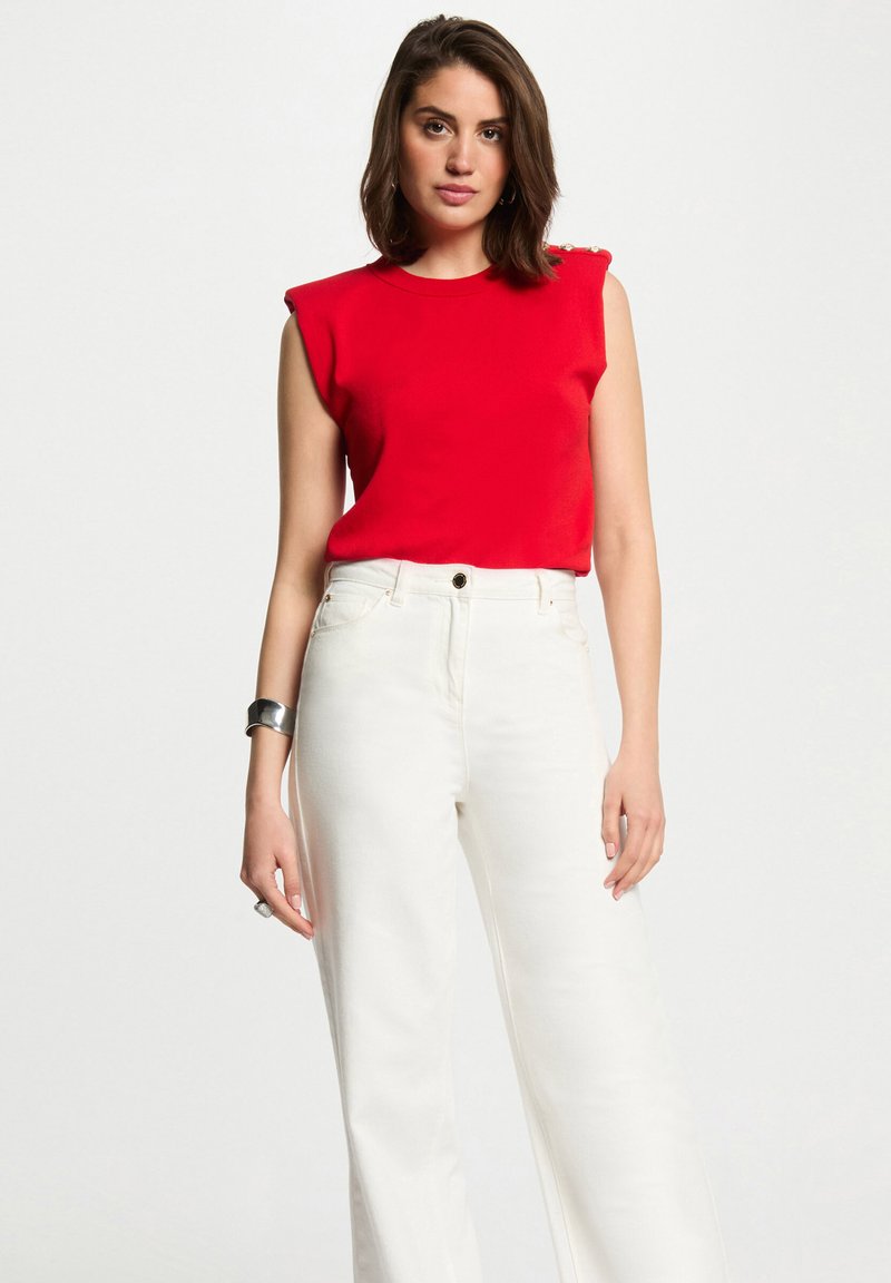 Top rouge sans manches avec rembourrage aux épaules, associé à un jean large taille haute blanc. Texture lisse en tricot et design ajusté.