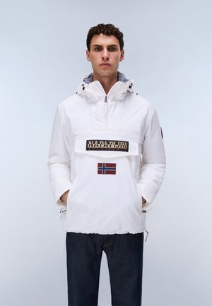 Mann trägt eine weiße Napapijri-Anorakjacke mit Kapuze und norwegischem Flaggenaufnäher, die Hände in den Taschen, vor einem schlichten, hellen Hintergrund stehend.